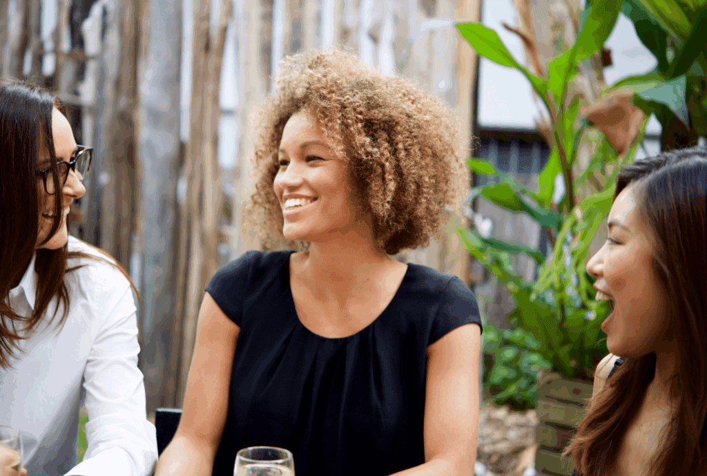 2 female friends laughing together. They are sat outside and have drinks with them.