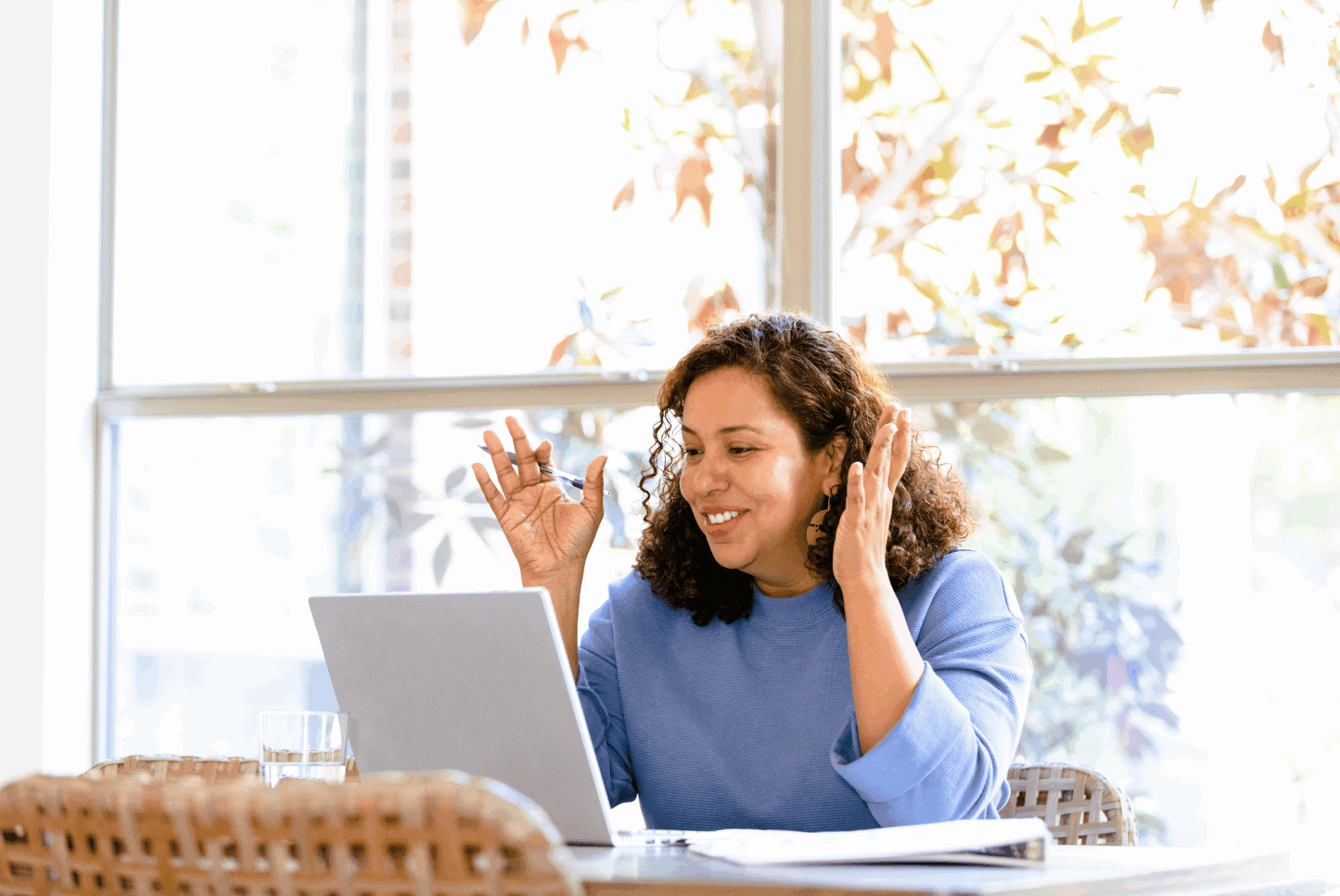 Female with curly hair sat at a desk in front of a window. She has a laptop on her desk and is talking to the person on the screen. She has her hands raised and has a pen in one hand