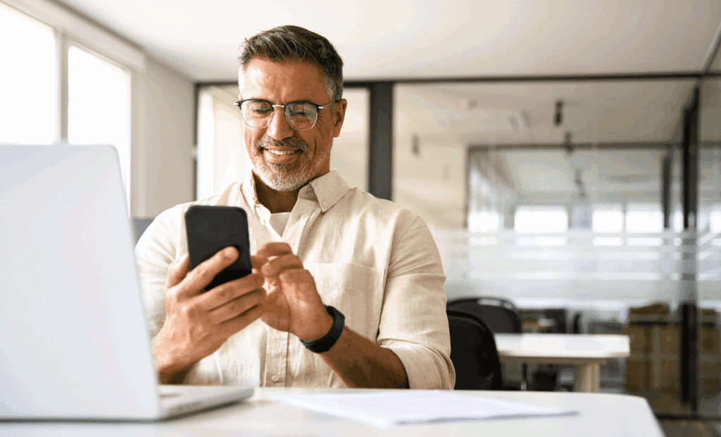 Mature male at office. He is sat at a desk with a laptop and papers on it. He has grey hair, wears glasses and has a beard. He wears a white shirt. He is holding a mobile phone and is looking at that