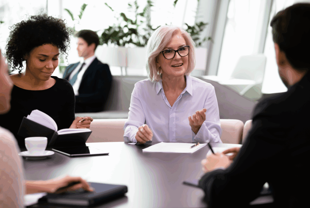 2 people at an office. We see the back of a head of a male, and a mature female talking to him. They both have pens and paper in front of them and a table between them.
