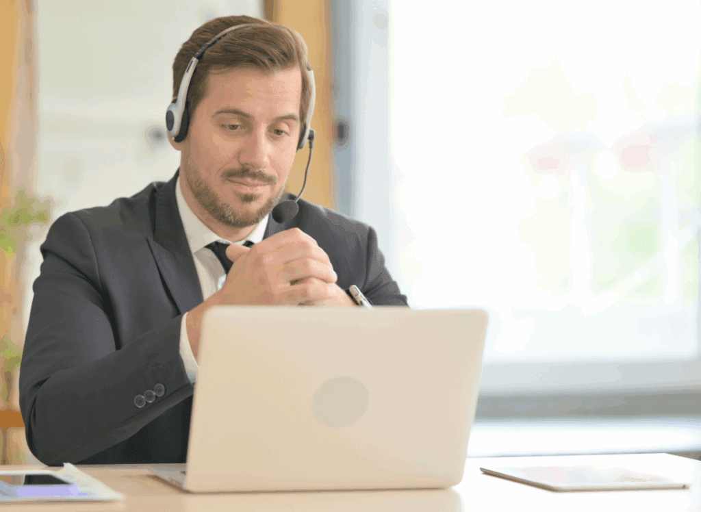 Male sat at desk. On the desk is a laptop which he is looking at. There are other things on his desk to the side. He is wearing a headset. He wears a grey suit