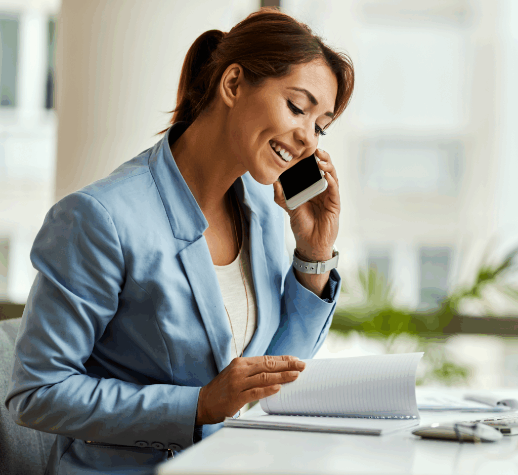Female sat at desk. She has a diary in front of her she is turning the pages in and she holds a mobile phone to her ear. She wears a pale suit and has her hair tied back.
