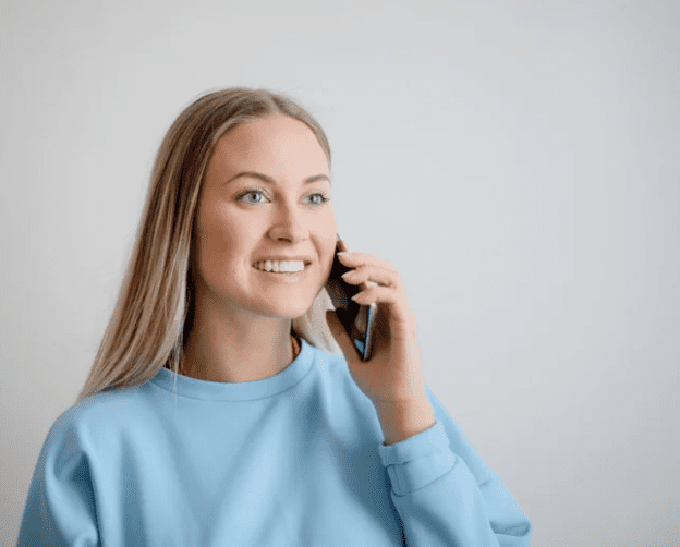 Female on phone call. She holds a mobile phone to her ear. She wears a pale blue sweater and has blonde hair. She is smiling