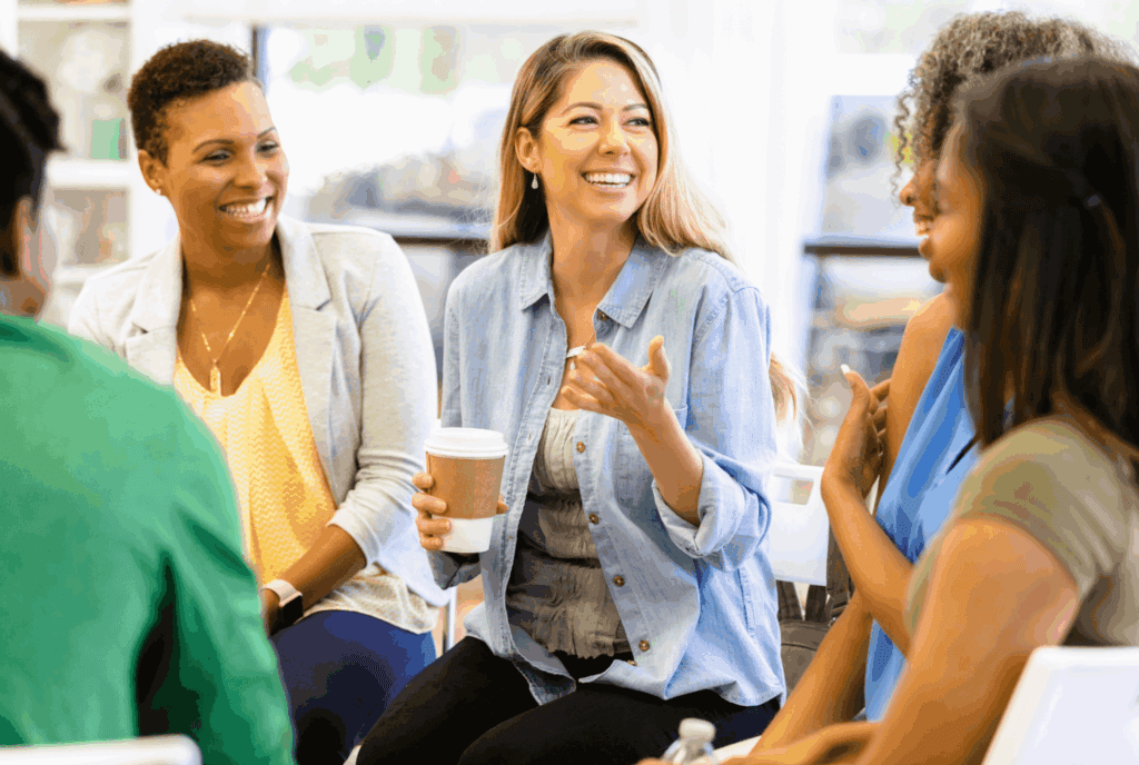 The photo shows 3 woman, who are in a casual setting talking. They are different ages and ethnicities. The seem to be sat in a coffee shop. One person smiles to the other two