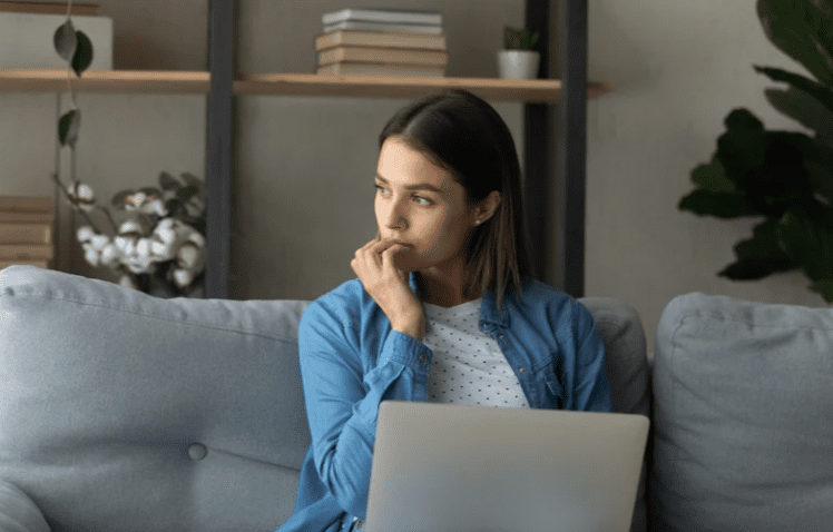 A woman in a blue shirt looking pensive while using her laptop on a comfortable sofa.