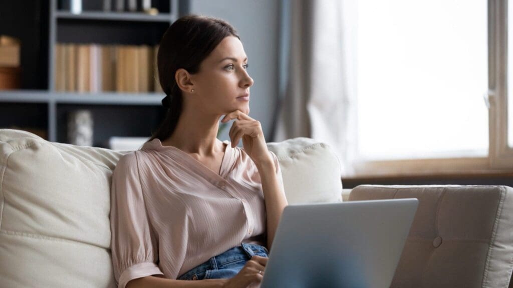 A woman sitting on a couch, engrossed in her laptop, seeking therapy for her low self-esteem.
