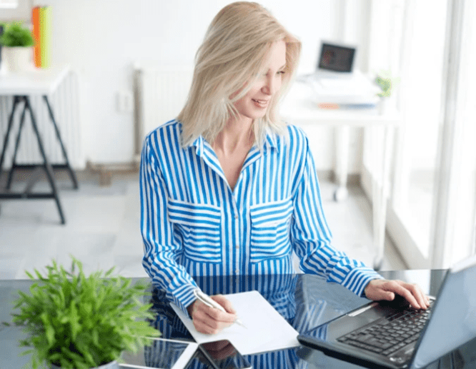 A woman in a blue striped shirt working at a desk with a laptop and notepad.