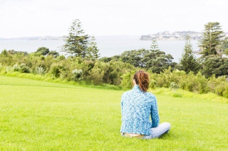A girl sitting on green grass, gazing at a scenic view of the sea and trees.