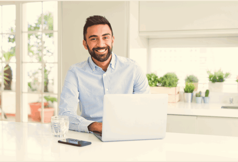 A smiling man in a striped shirt sitting at a desk with a laptop.
