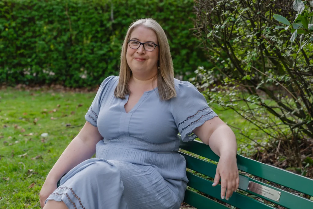 Hannah Paskin smiling while seated on a green bench in a garden, wearing a light blue outfit.