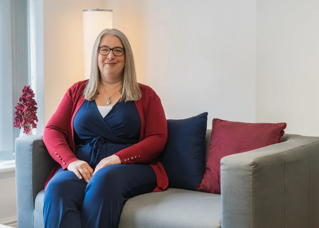 Hannah Paskin in a home office setting, smiling while seated on a gray sofa with decorative cushions.