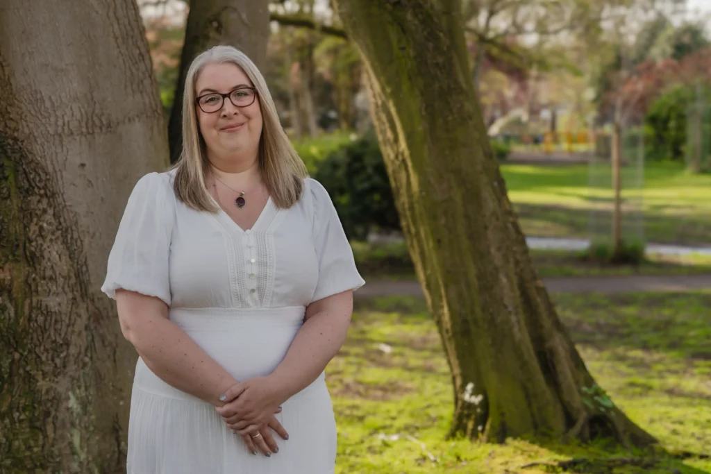 A woman in a white dress standing in a park with trees in the background.