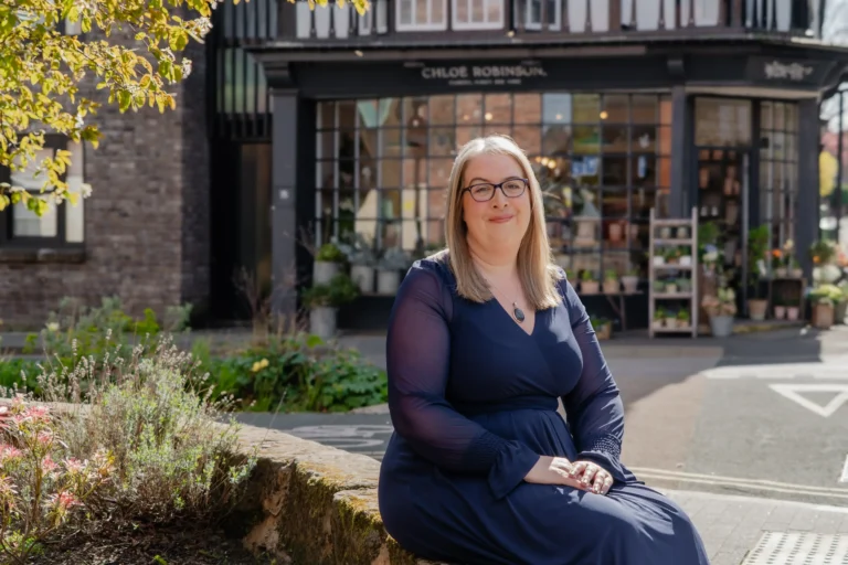 Hannah Paskin with glasses in a navy dress sitting on a stone wall outside a boutique.